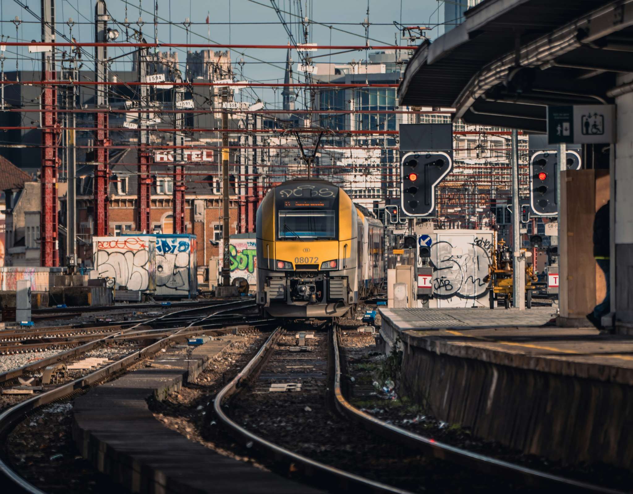 Frontale aanblik van een gele SNCB-trein op druk spoorwegknooppunt met seinen.
