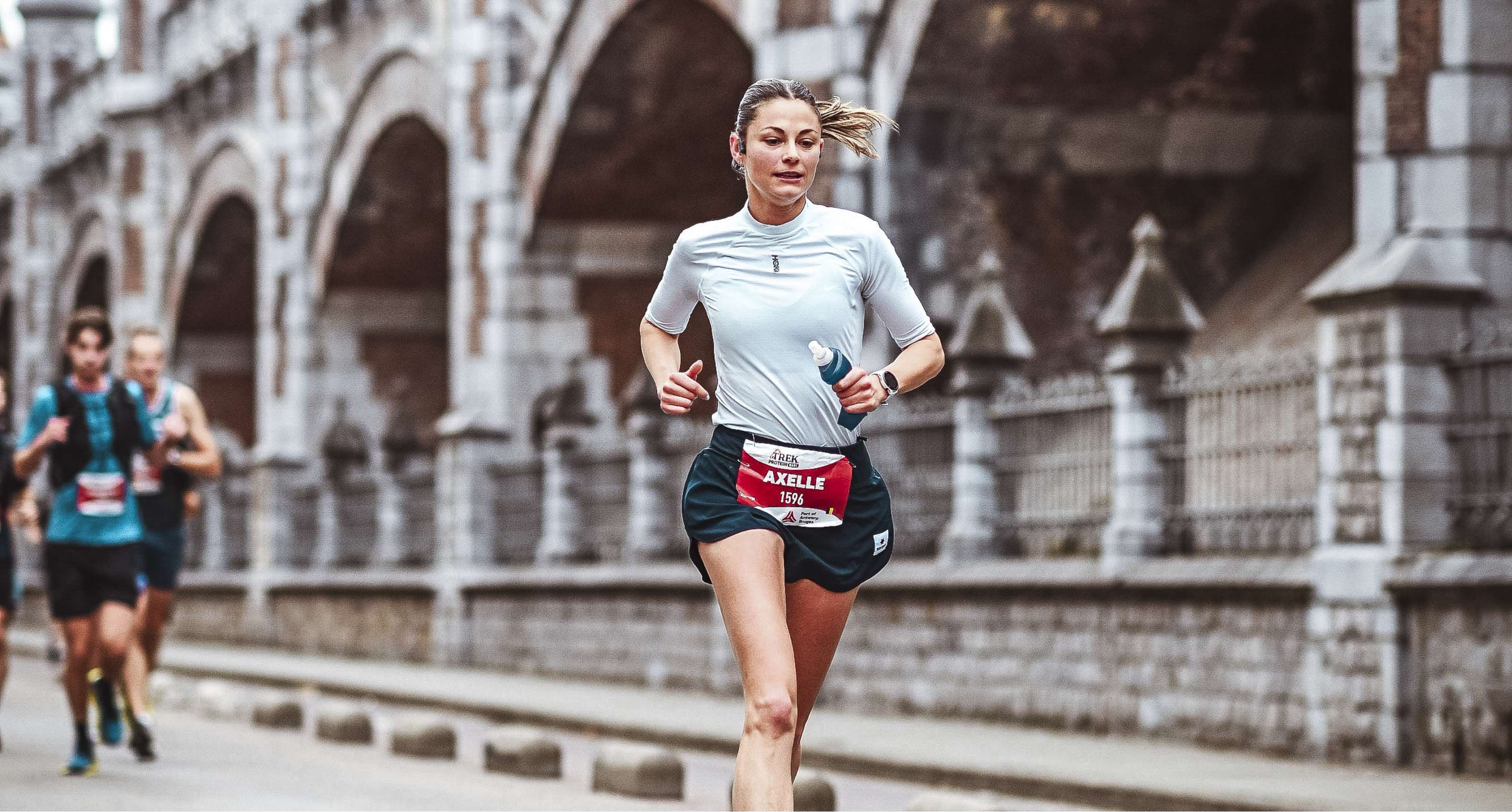 Axelle loopt met startnummer 1596 tijdens een hardloopwedstrijd langs historische bogen.