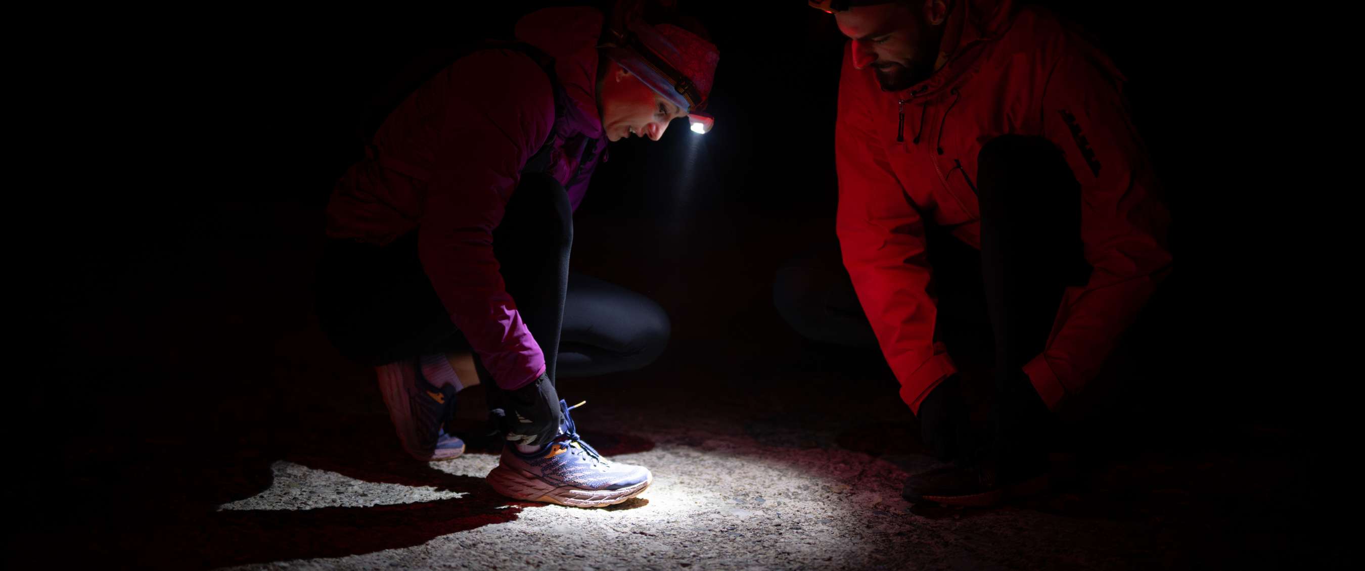 Twee lopers hurken in het donker terwijl één zijn schoenveters strikt, verlicht door een hoofdlamp.