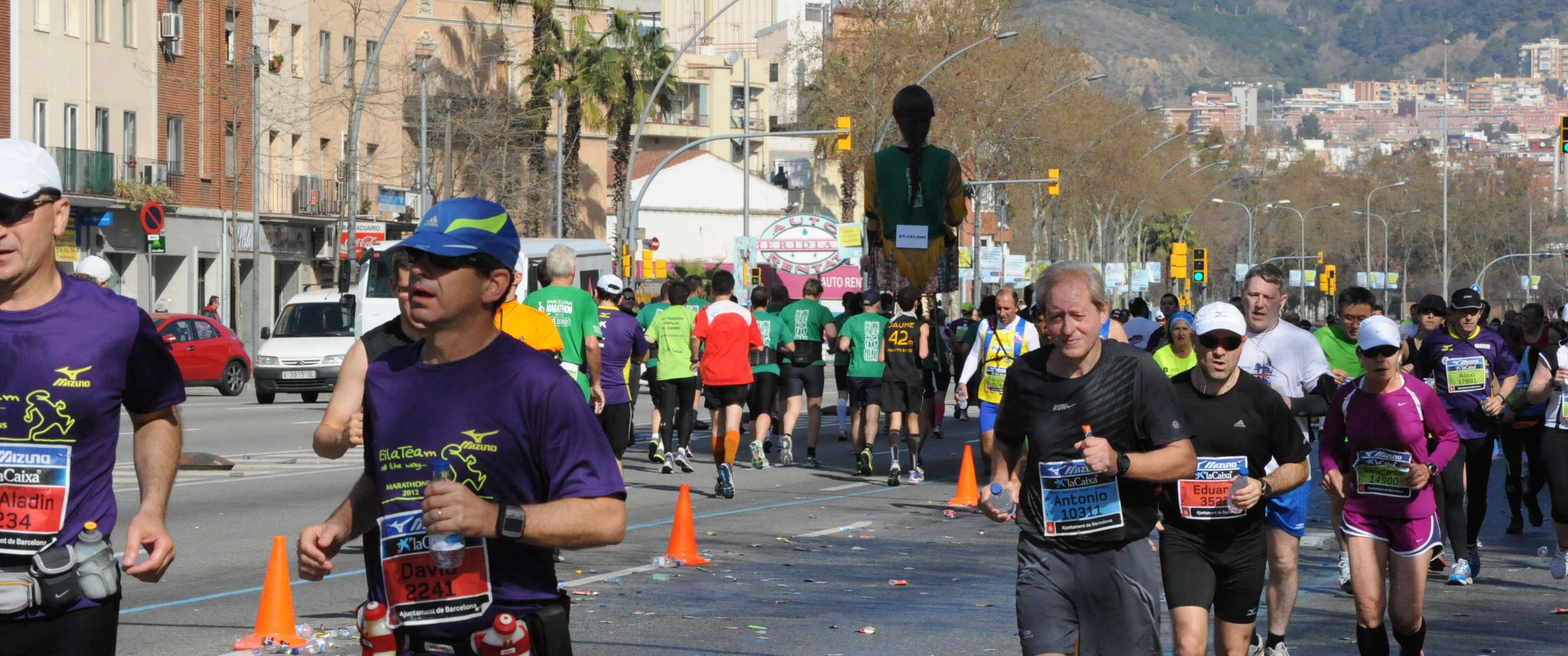Groep hardlopers tijdens een stadsmarathon in Barcelona, met gebouwen, palmbomen en heuvels op de achtergrond