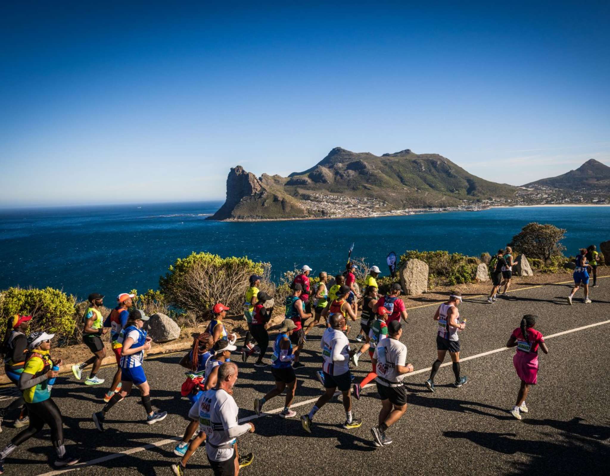 Marathonlopers rennen over een kustweg met uitzicht op zee en bergen op de achtergrond