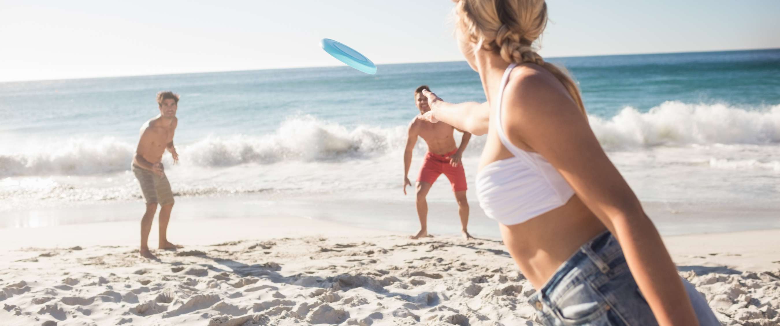 Drie personen spelen met een blauwe frisbee op het strand bij de branding.
