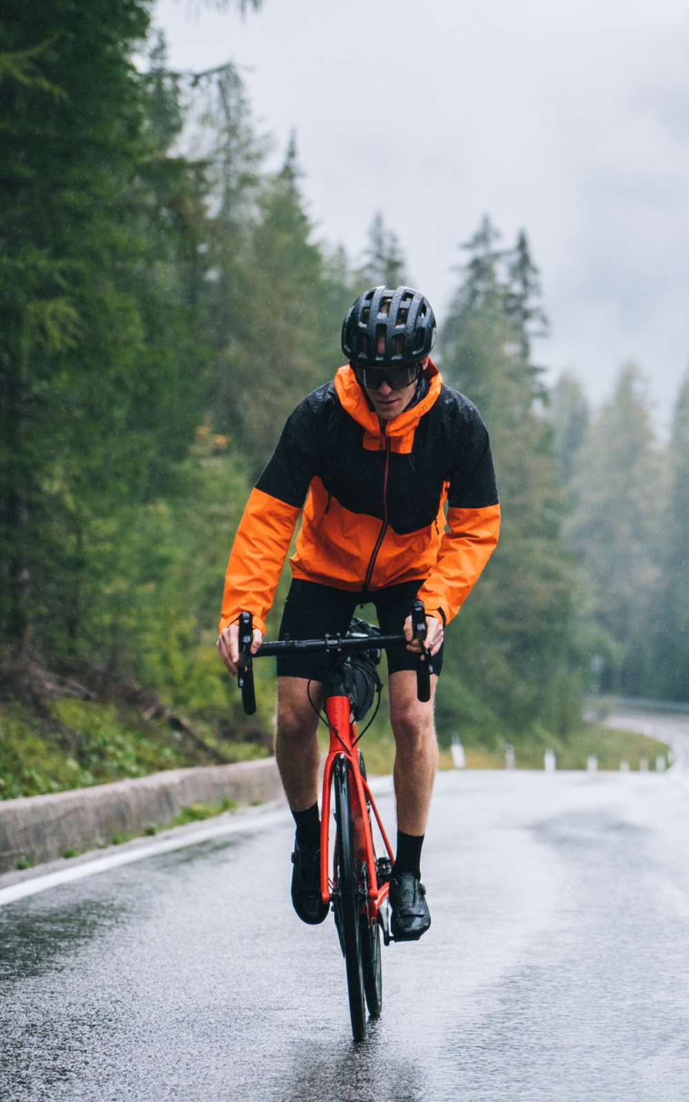 Wielrenner in zwart-oranje regenjas rijdt door de regen over een natte weg langs naaldbomen.