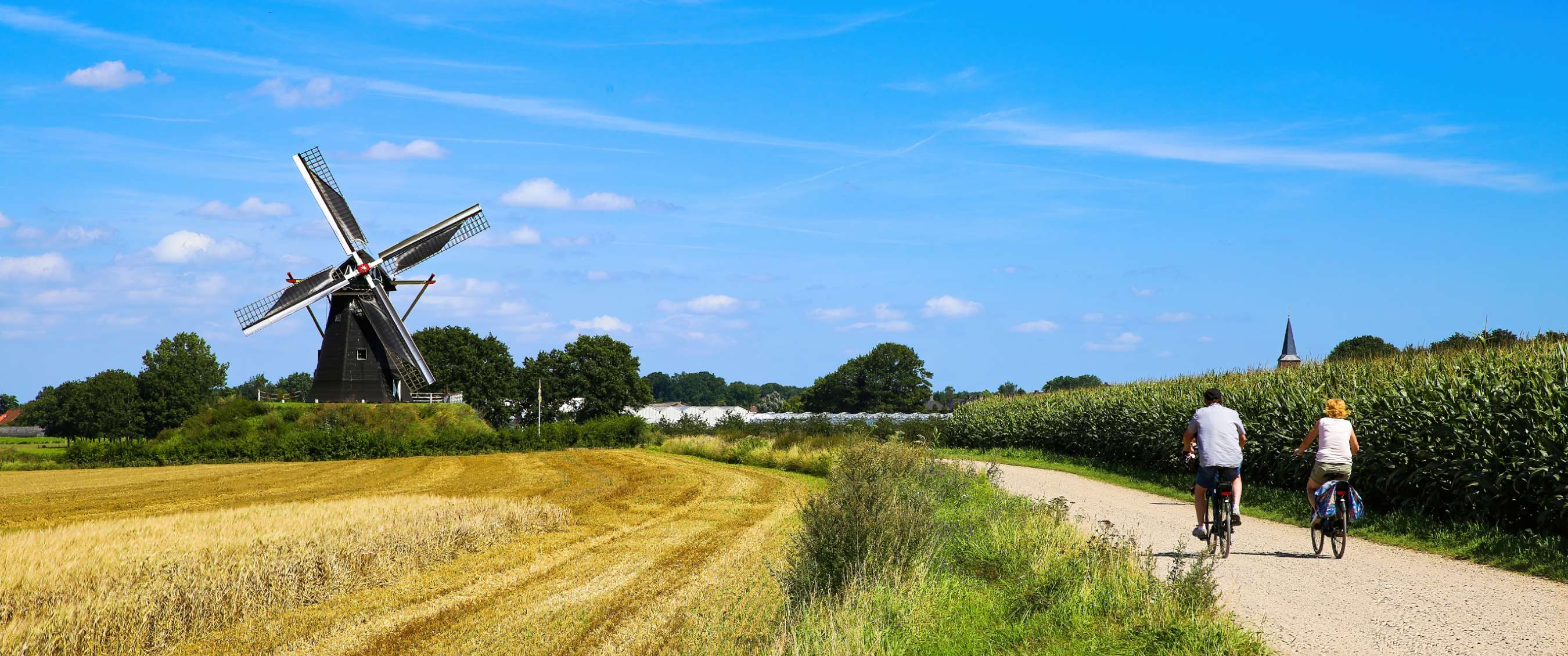 Landelijk fietspad met fietser, windmolen, akkers en blauwe lucht.