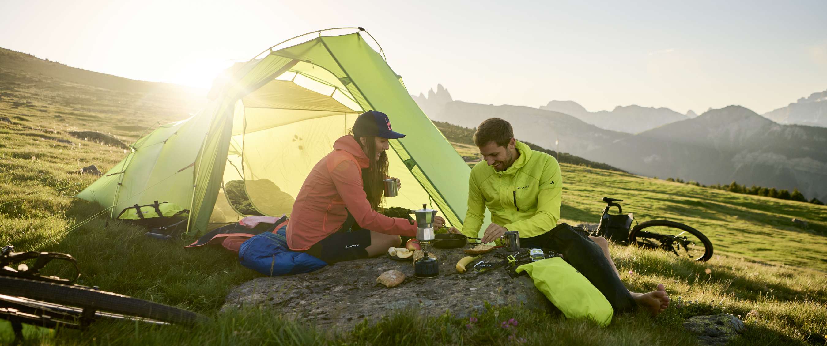 Twee personen koken bij een groene tent op een bergweide met fietsen naast de kampeerplek.