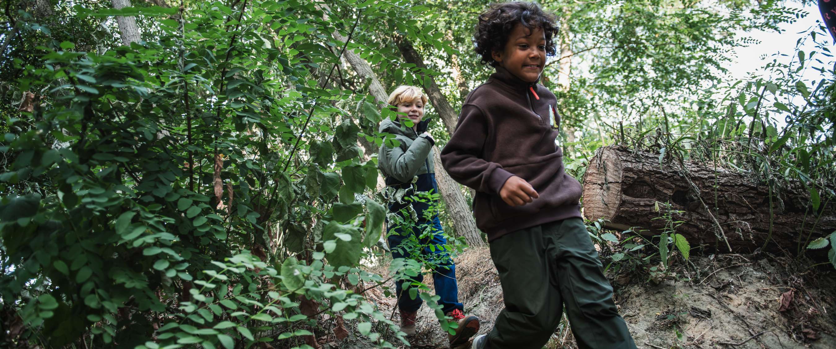 Twee kinderen in buitenkleding volgen een smal pad tussen groene begroeiing en boomstammen.