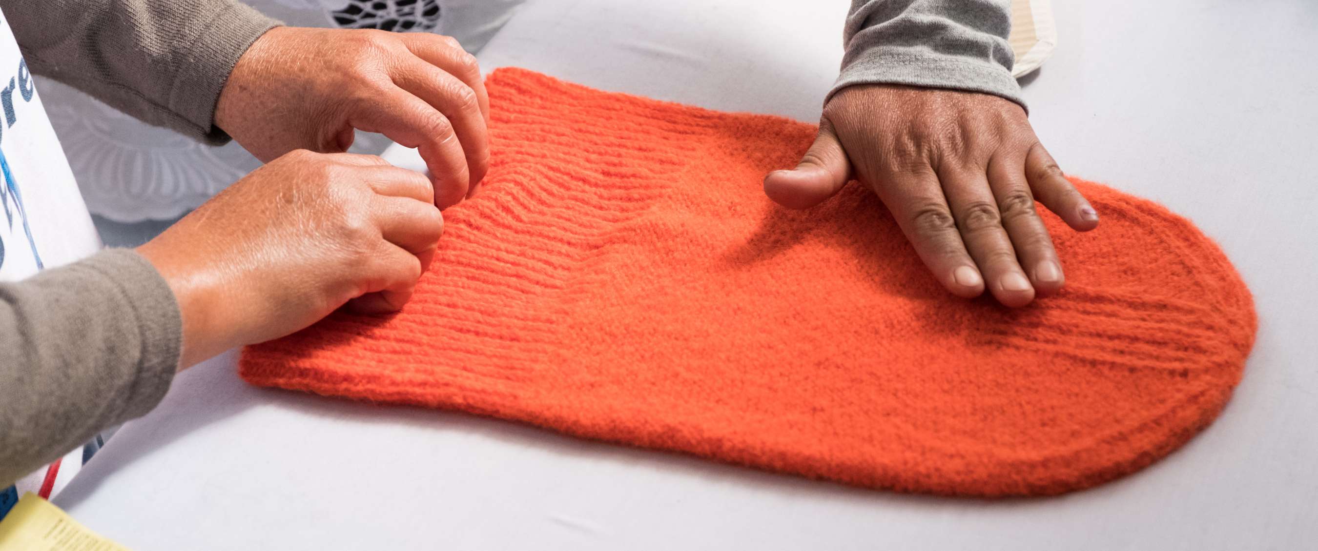 Twee paar handen onderzoeken een feloranje gebreid kledingstuk op een tafel.