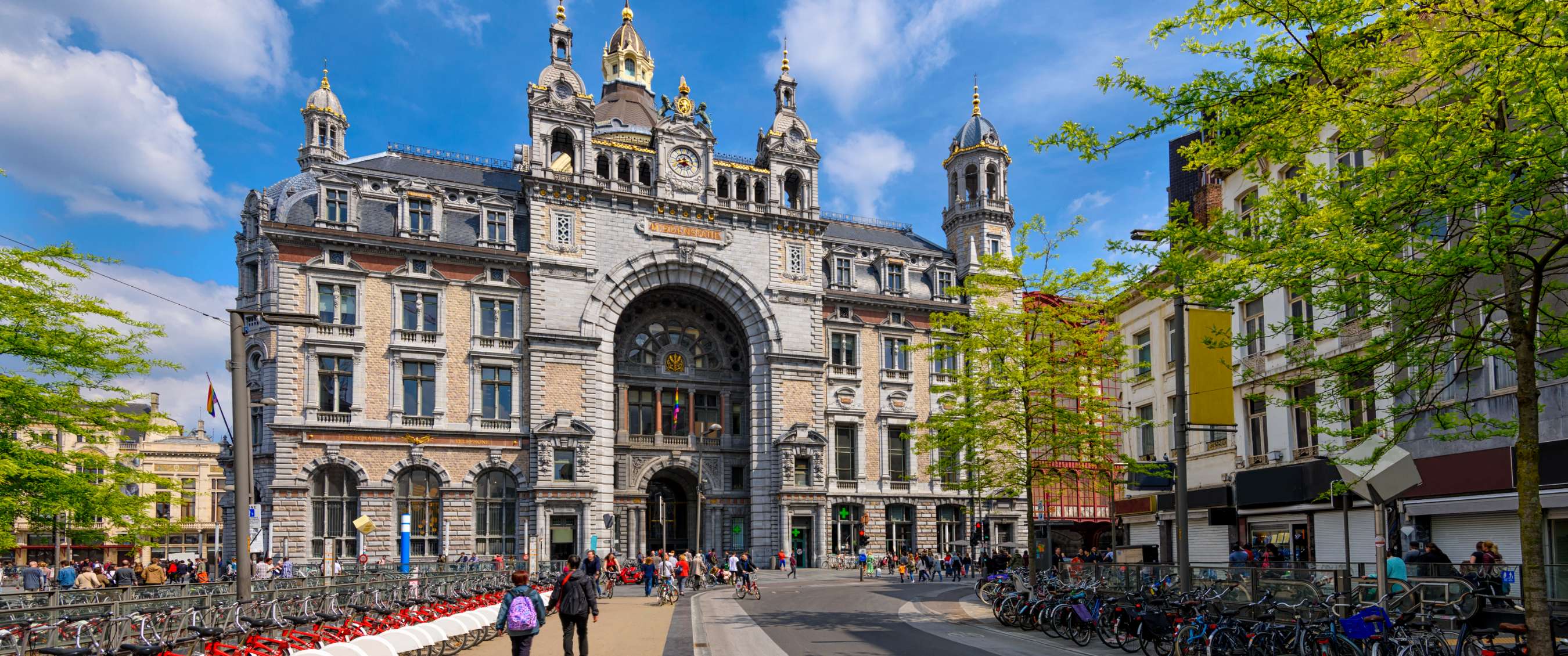 Grote historische stationsgevel met torens, fietsen en voorbijgangers onder een blauwe lucht.