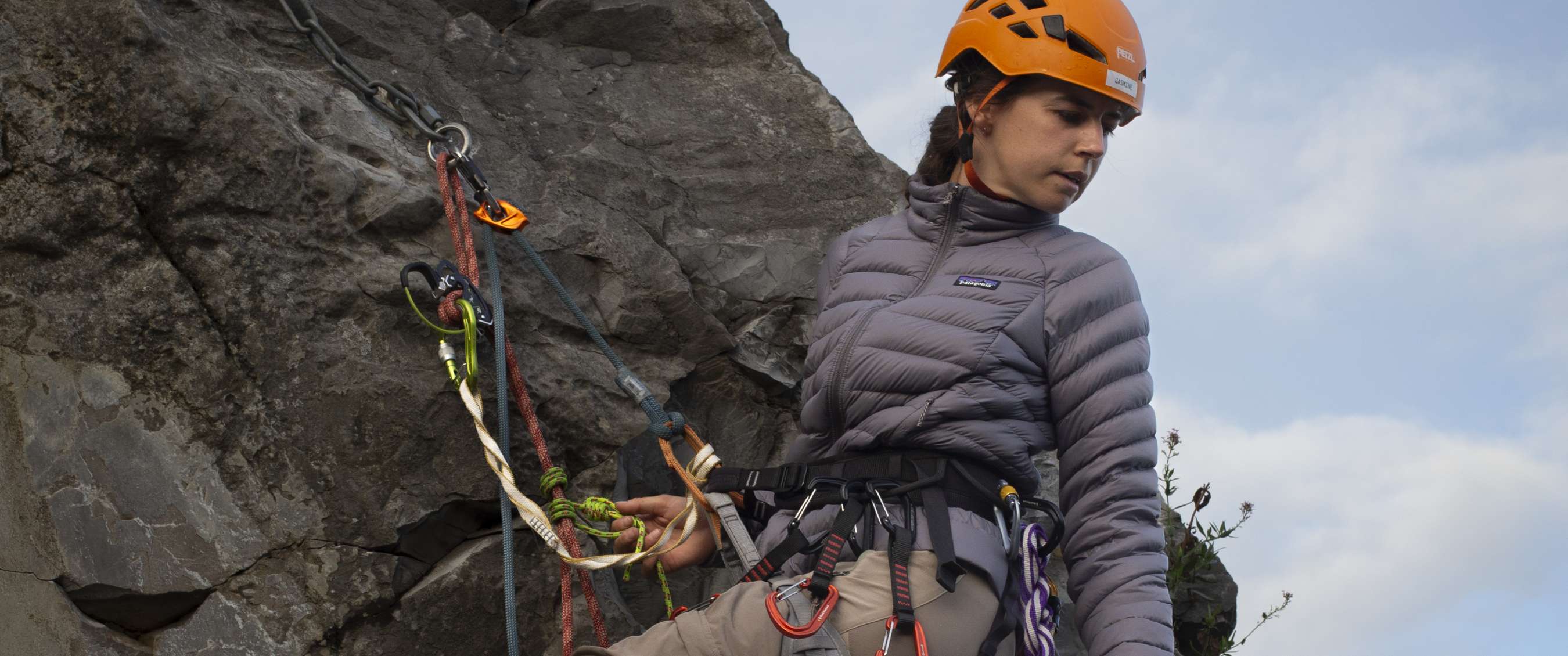 Klimmer met oranje helm en grijze Patagonia-jas staat gezekerd aan een steile rotswand.