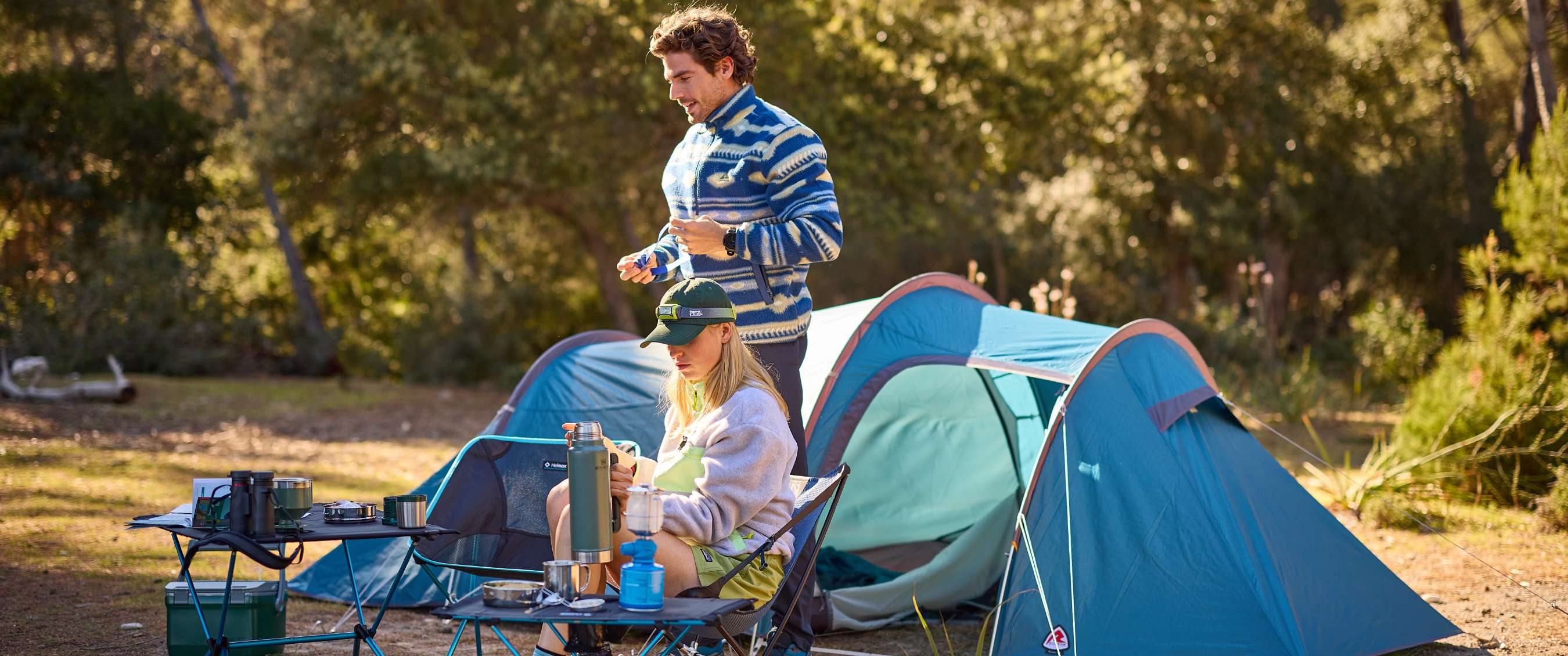 wee personen bij blauwe tenten met campingstoelen, tafel en kookgerei.
