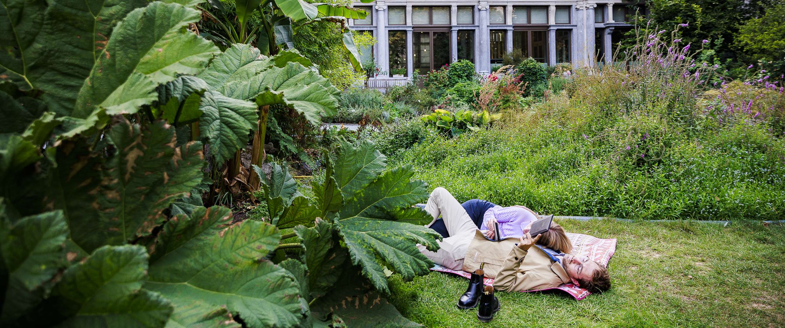 Twee personen liggen op de grasmat in een groen stadspark met historisch gebouw op achtergrond.