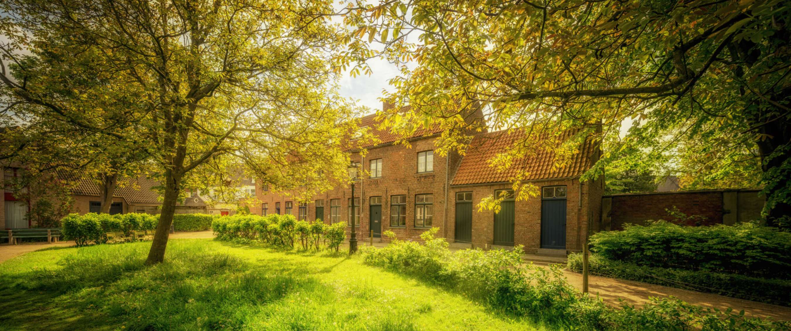 Zonlicht valt op historische bakstenen huizen rond groen binnenhof in Brugge.