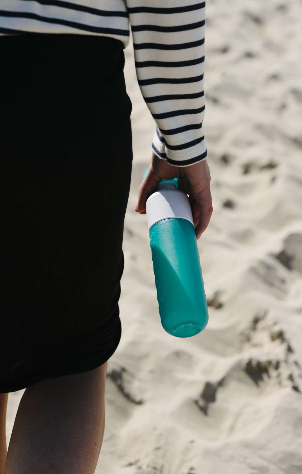 Persoon met gestreepte trui draagt een turquoise drinkfles op zand