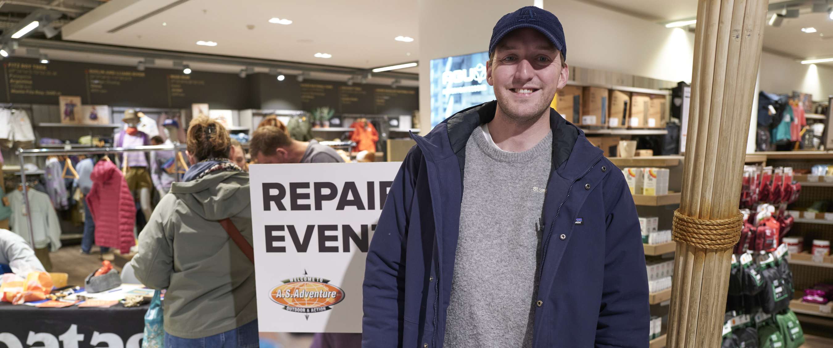 Man in blauwe jas staat in een winkel voor een bord met tekst REPAIR EVENT.