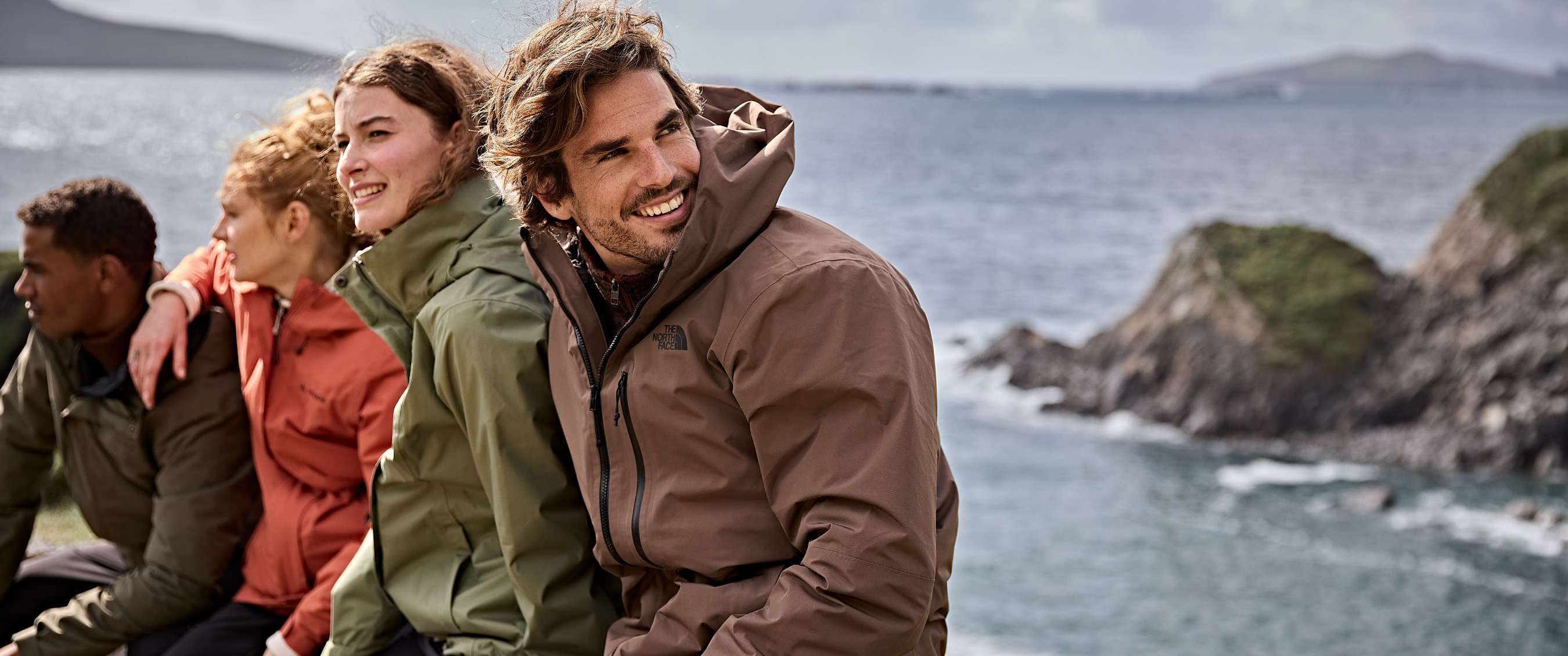 Twee vrouwen en twee mannen in gekleurde jassen ontspannen aan zee met heuvels op de achtergrond.