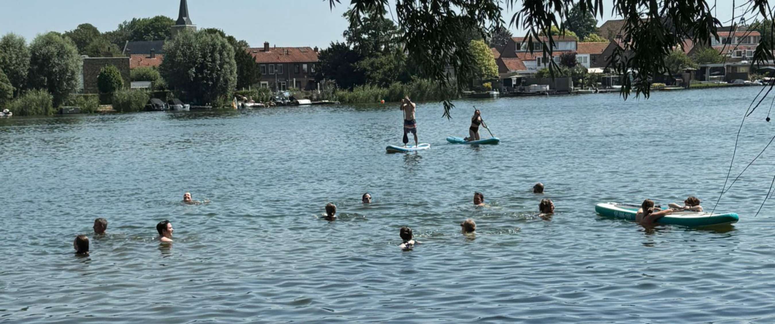 Zwemmers en twee paddleboarders bevinden zich op rustig water bij een bebouwde oever.