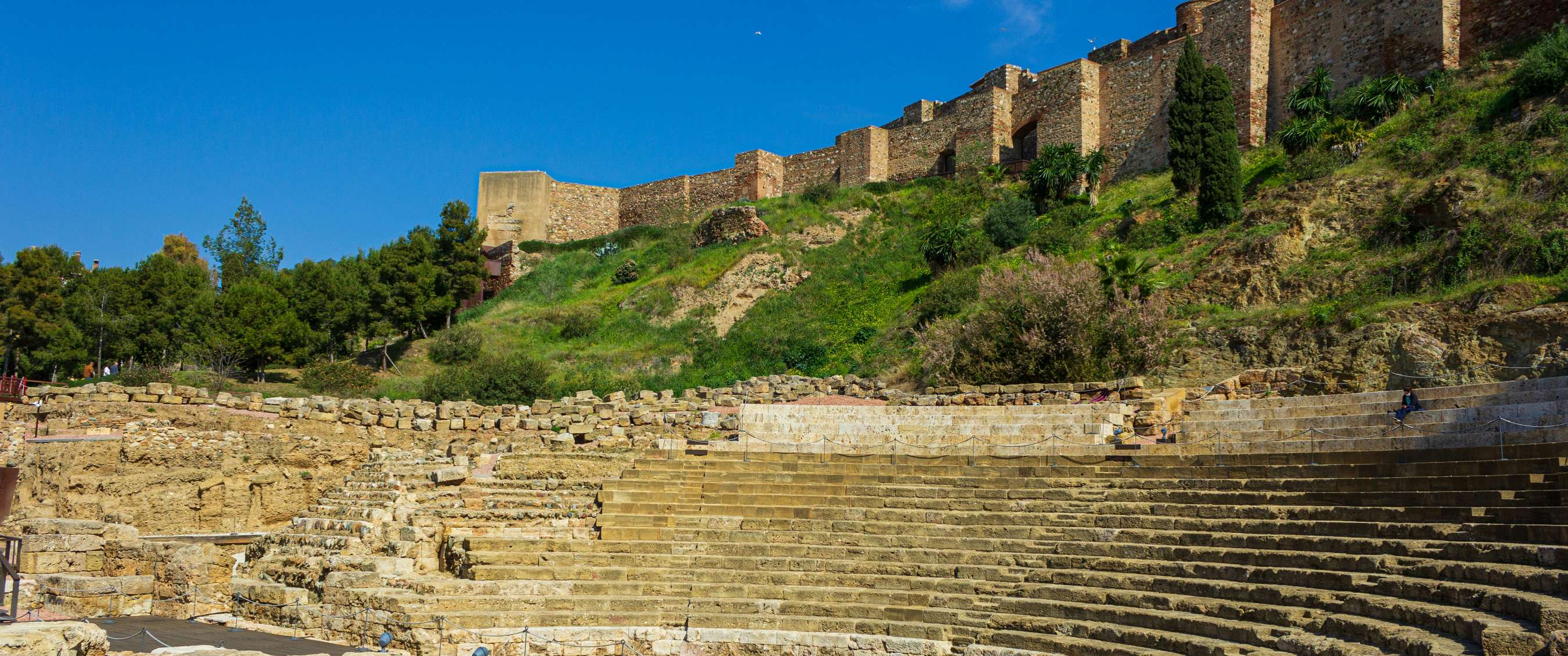 Romeins theater met stenen zitrijen en vestingmuren op een groene heuvel in Málaga.