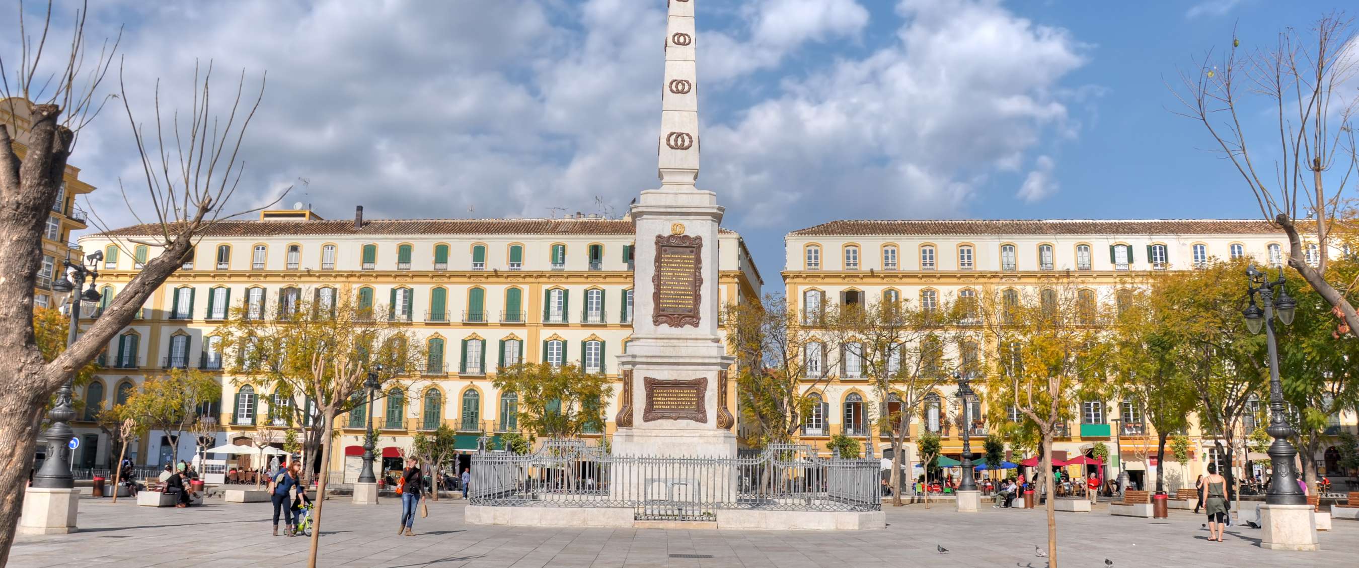 Witte obelisk op een ruim stadsplein in Málaga, omringd door historische gevels en bomen.