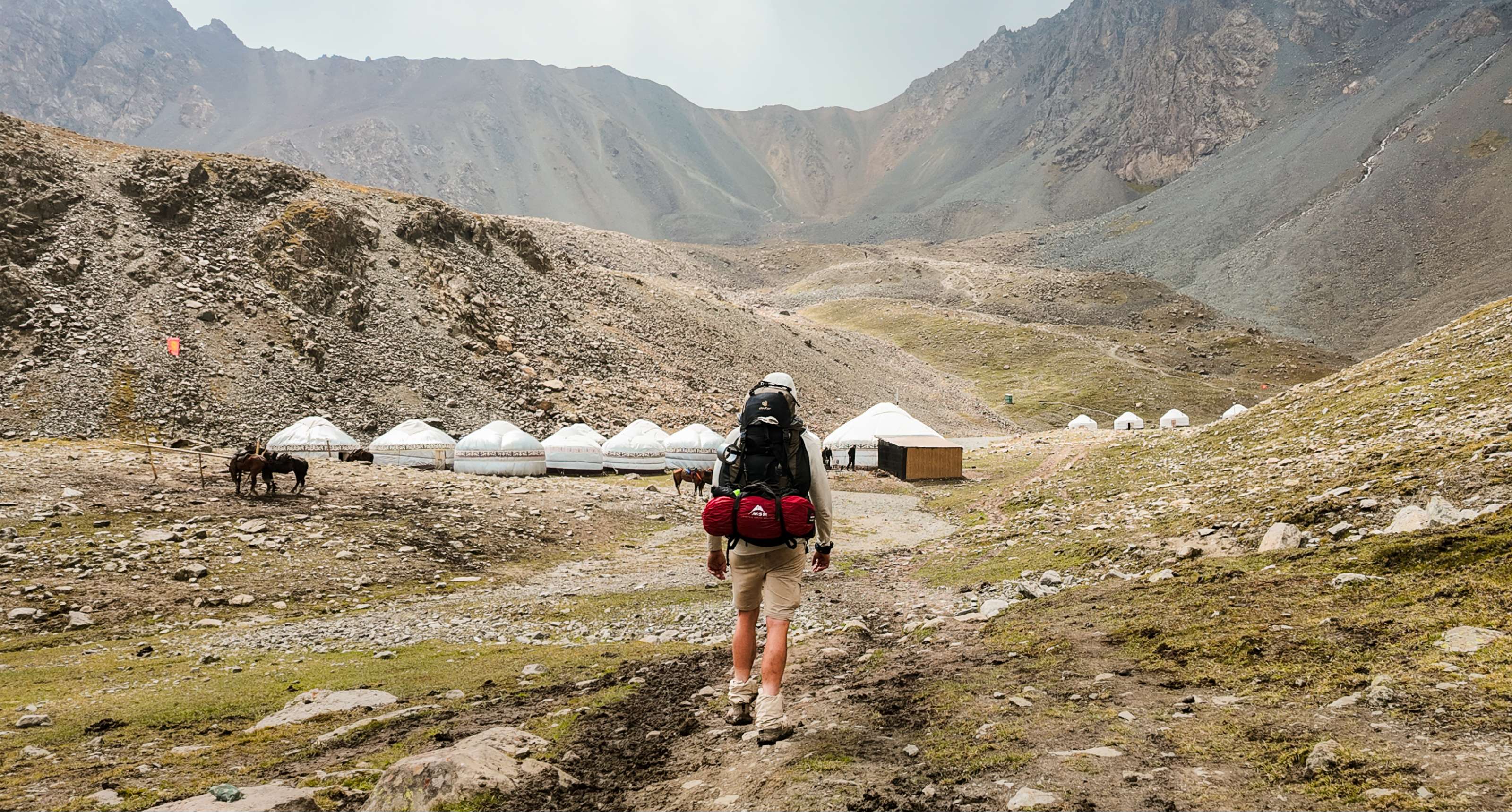 Backpacker wandelt over rotsachtig pad naar witte yurts in een afgelegen bergkamp.