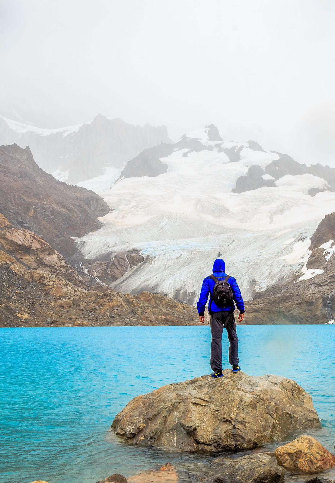 Wandelaar met blauwe jas kijkt uit over helderblauw bergmeer en besneeuwde gletsjer.