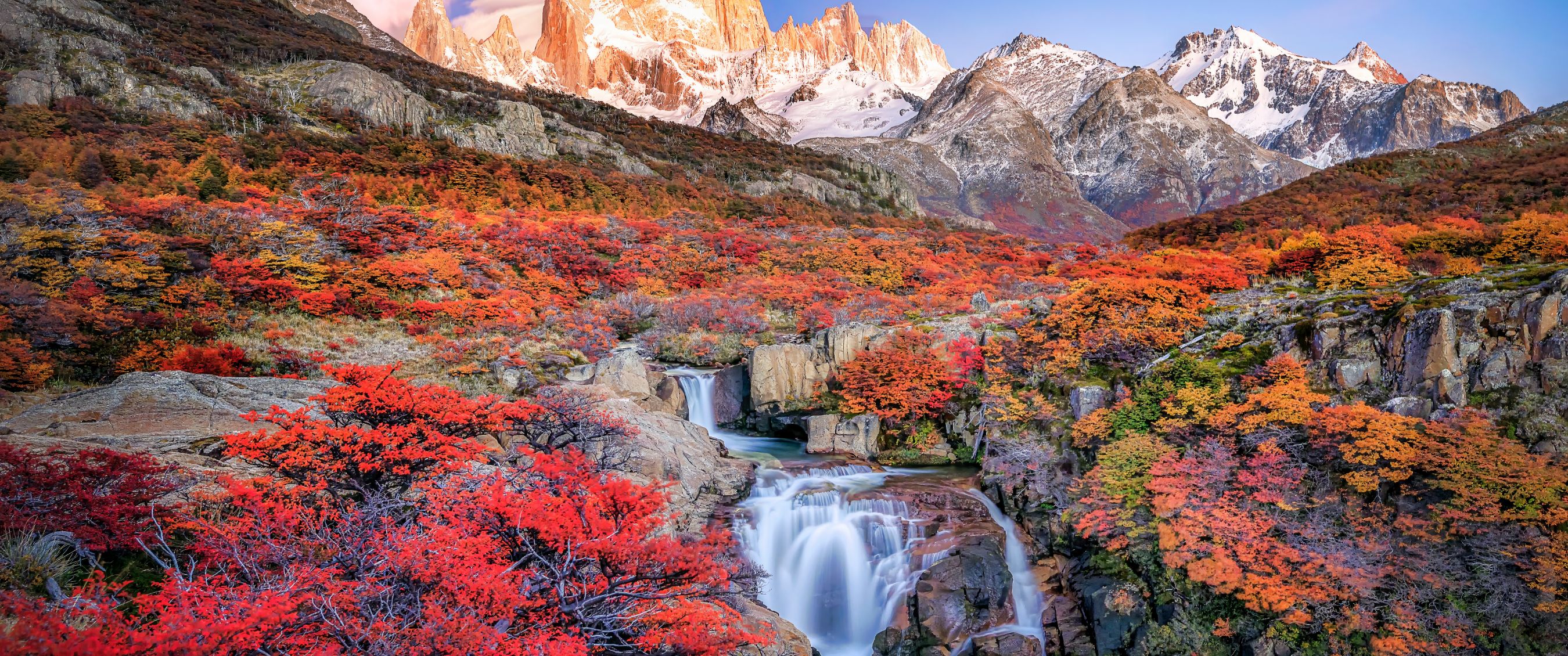 Waterval tussen felgekleurde herfstbomen met besneeuwde bergtoppen op de achtergrond.
