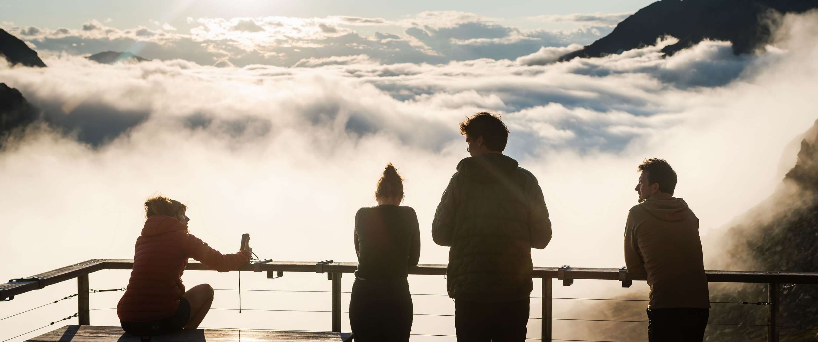 Vier personen staan aan een uitkijkplatform boven een wolkenlaag.