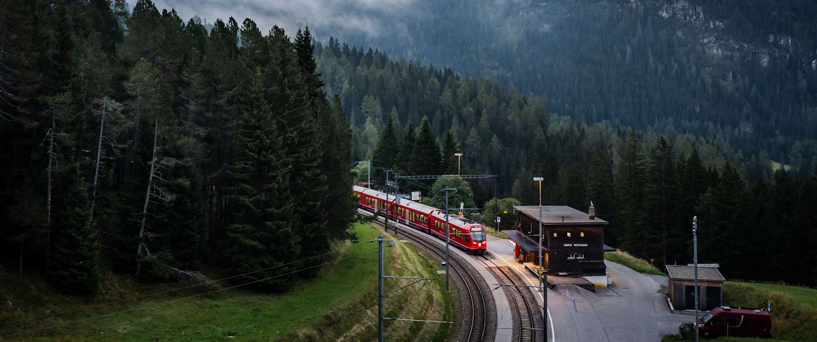 Smalspoortrein rijdt langs een bergstation omringd door naaldbomen.