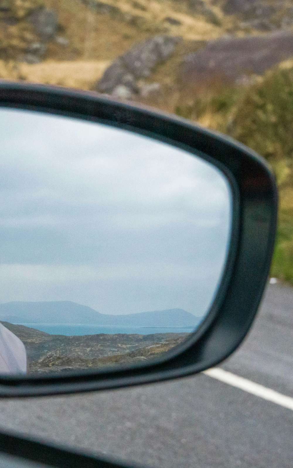 Autospiegel met scherp zicht op kust, zee en heuvels langs een bergweg.