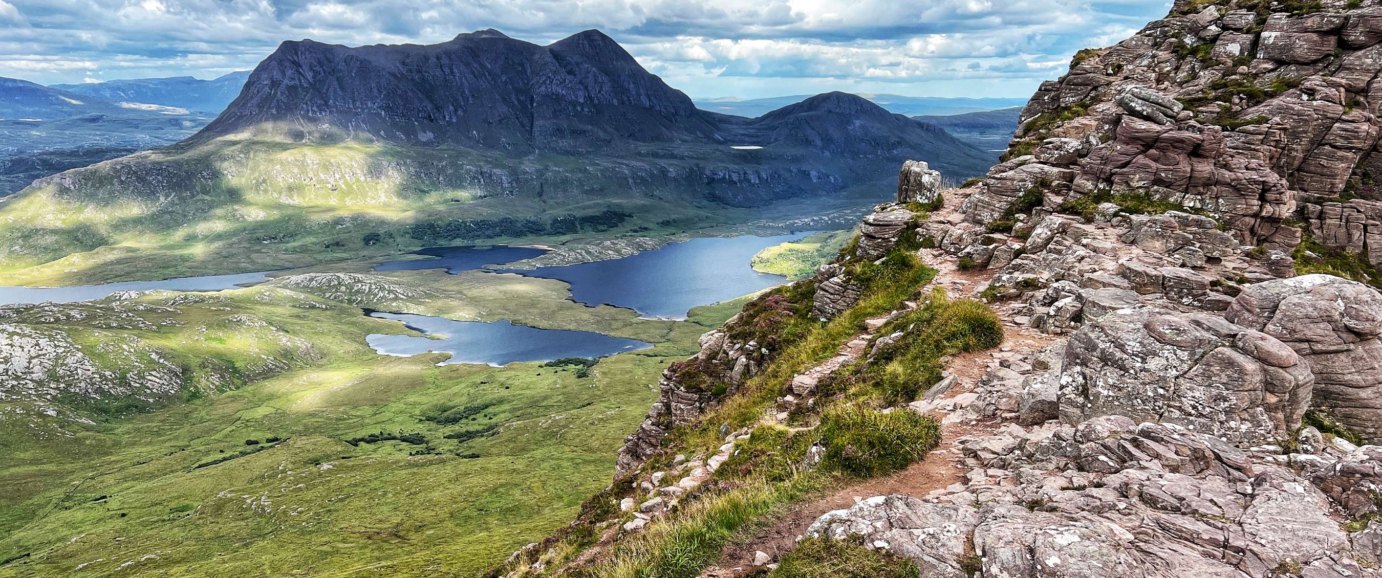 Uitzicht vanaf een rotsachtig bergpad op groene valleien, donkere bergen en meerdere meren.
