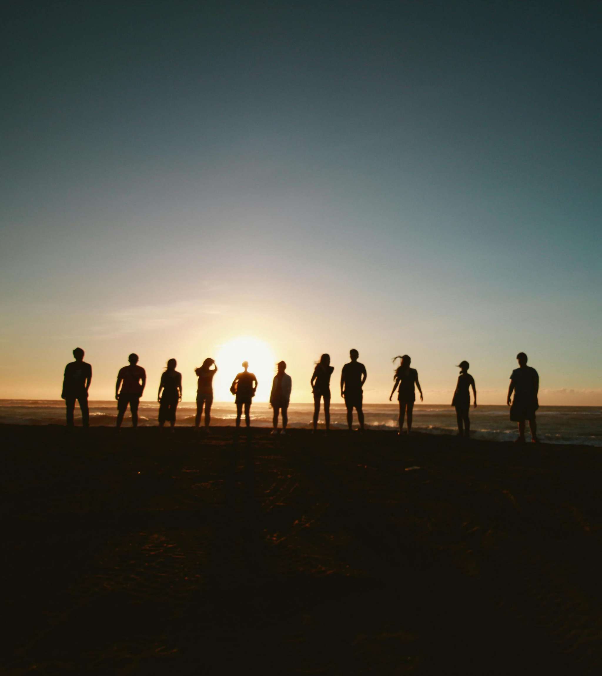 Groep mensen als silhouetten op het strand met de zon laag aan de horizon.