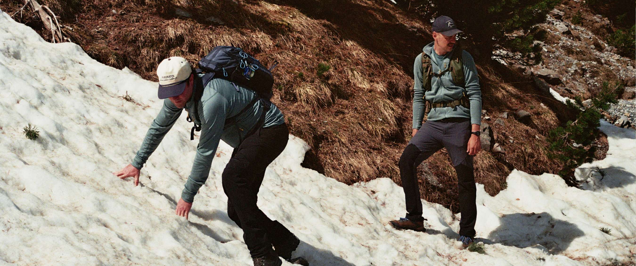 Twee wandelaars staan bij een bergmeer met besneeuwde rotsachtige bergen op de achtergrond.