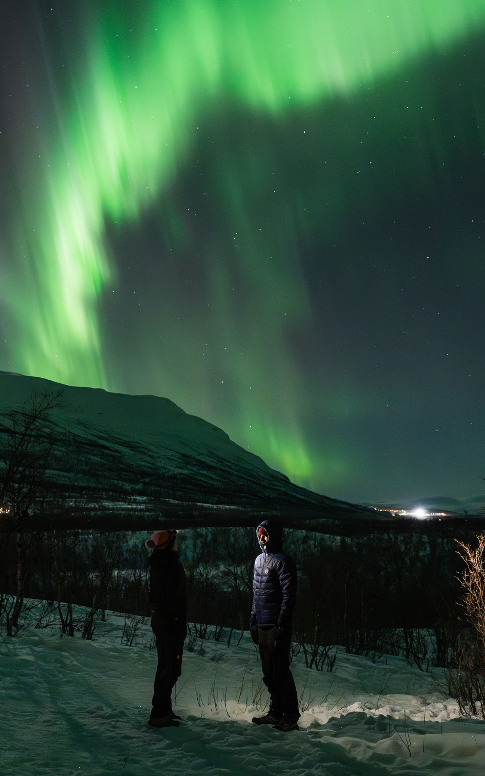 Groen noorderlicht boven besneeuwde bergen met twee personen in het donker.