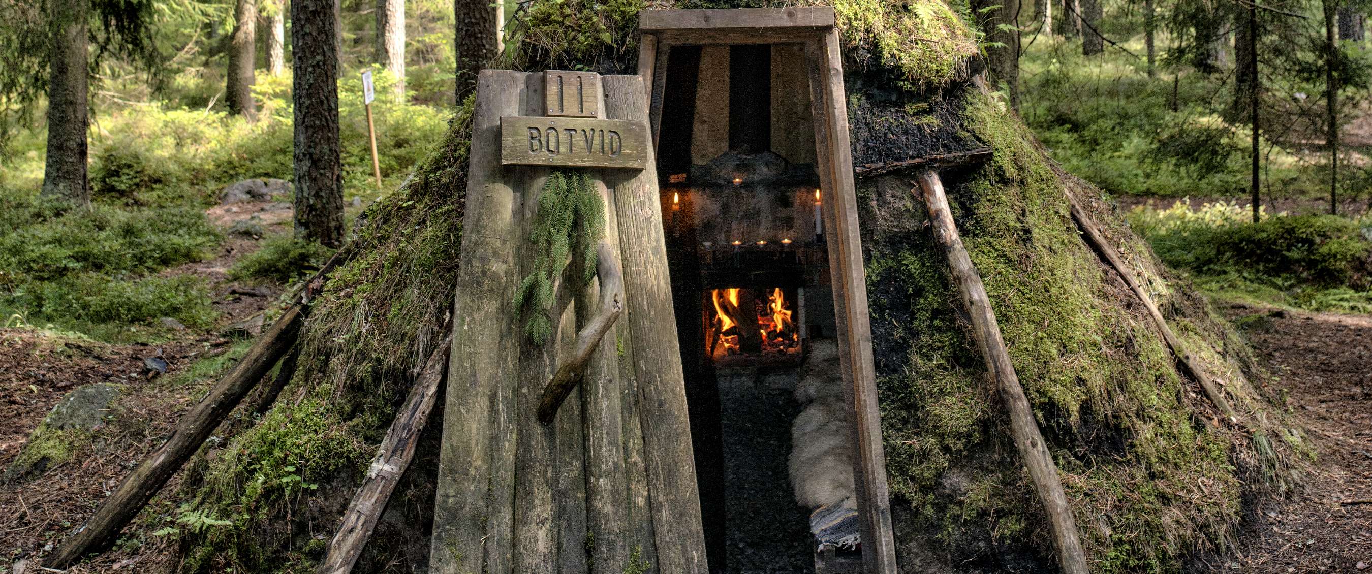 Houten bosverblijf bedekt met mos, met open deur, vuurplaats binnen en bord met tekst “BOTVID”.