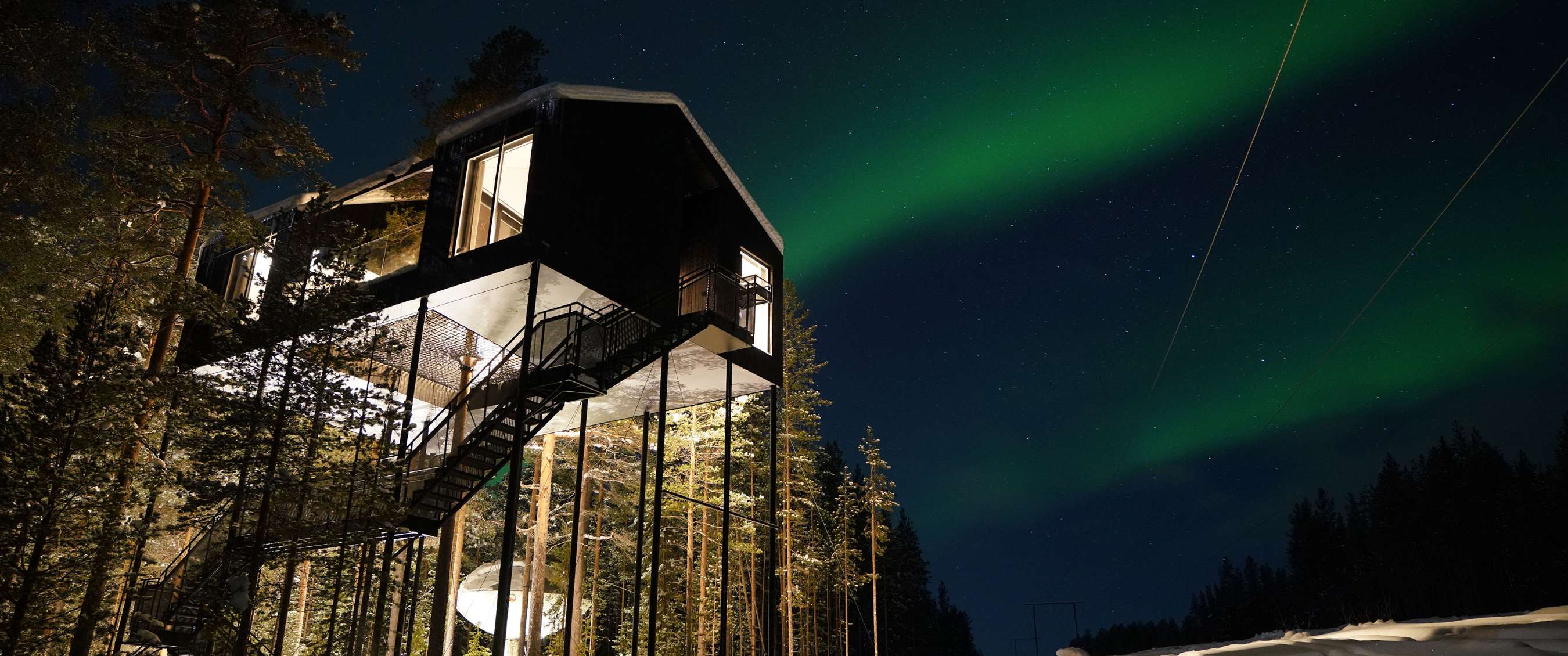 Houten boomhut op palen in een besneeuwd bos, verlicht interieur en aurora borealis op de achtergrond.