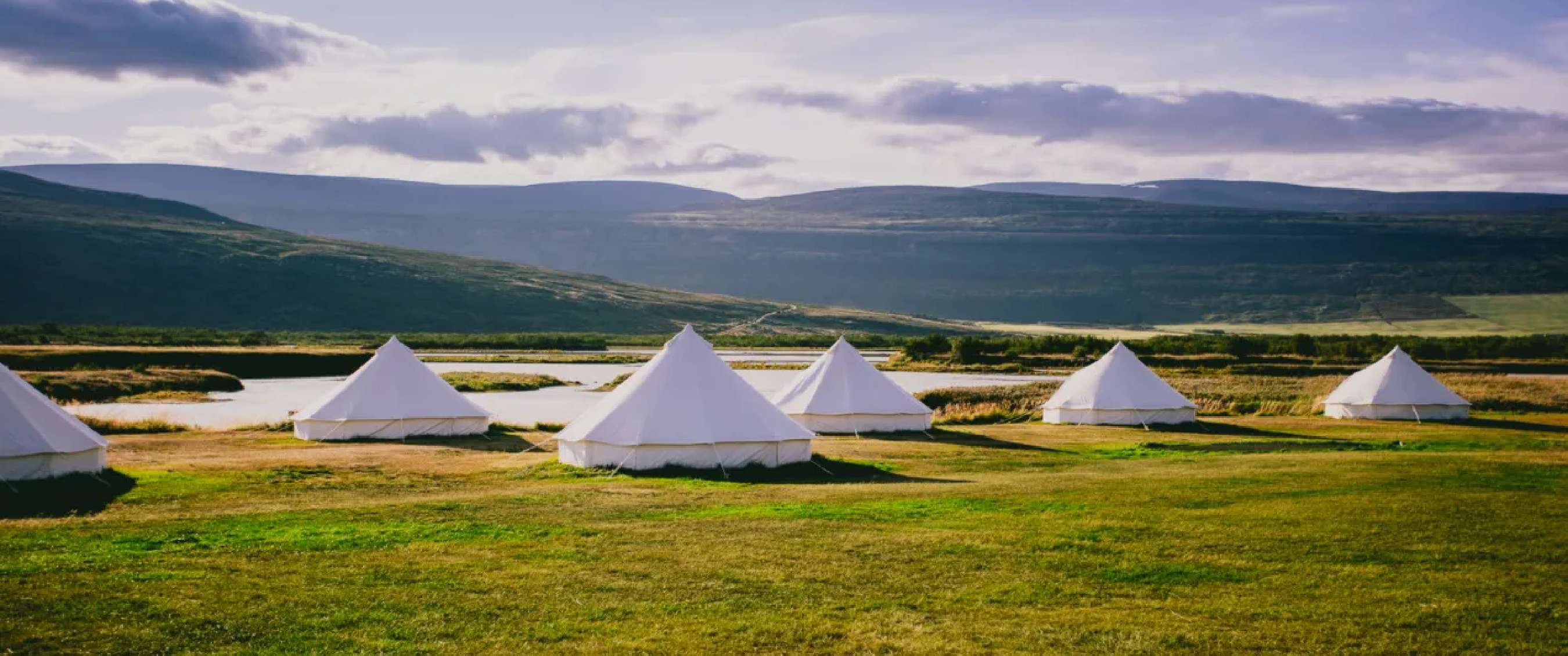 Witte glampingtenten opgesteld in een open graslandschap met rivier en bergen op de achtergrond.