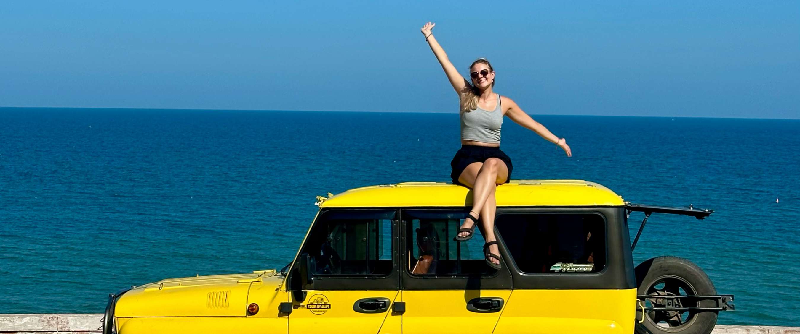 Persoon zit zwaaiend op gele jeep met blauwe zee op de achtergrond