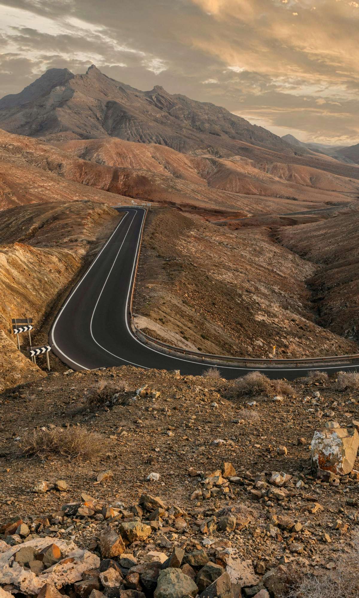 Slingerende asfaltweg door een droog berglandschap bij zonsondergang.