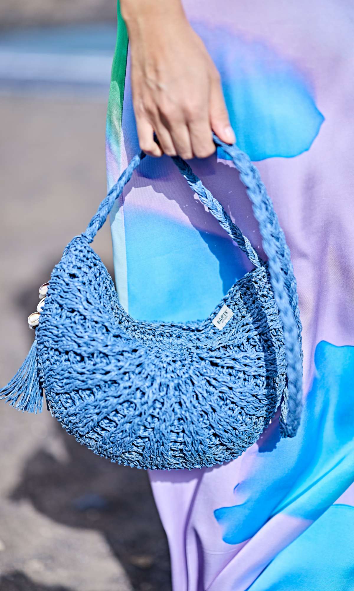 Hand die een blauwe gehaakte handtas vasthoudt voor een paarse jurk met blauwe print.