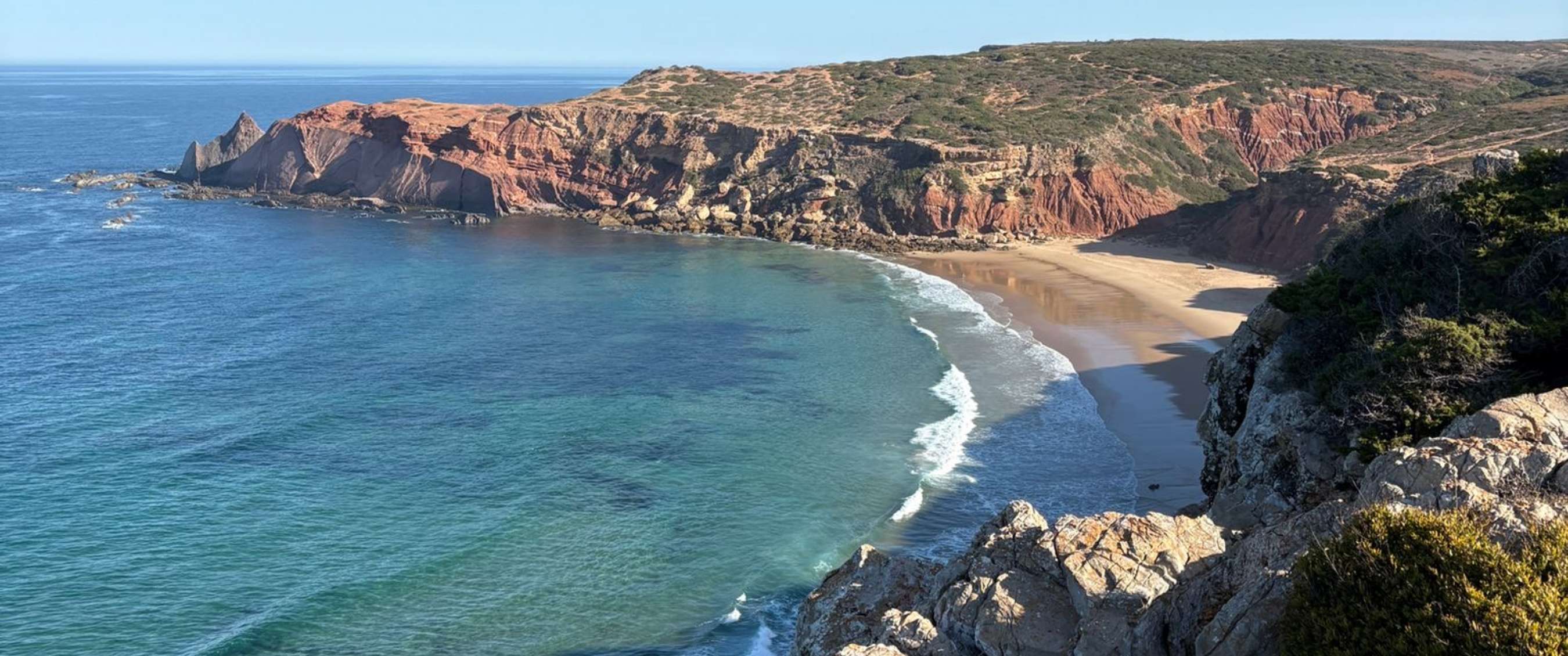 Uitzicht op rotsachtige kust, zandstrand en blauwe oceaan onder heldere lucht.