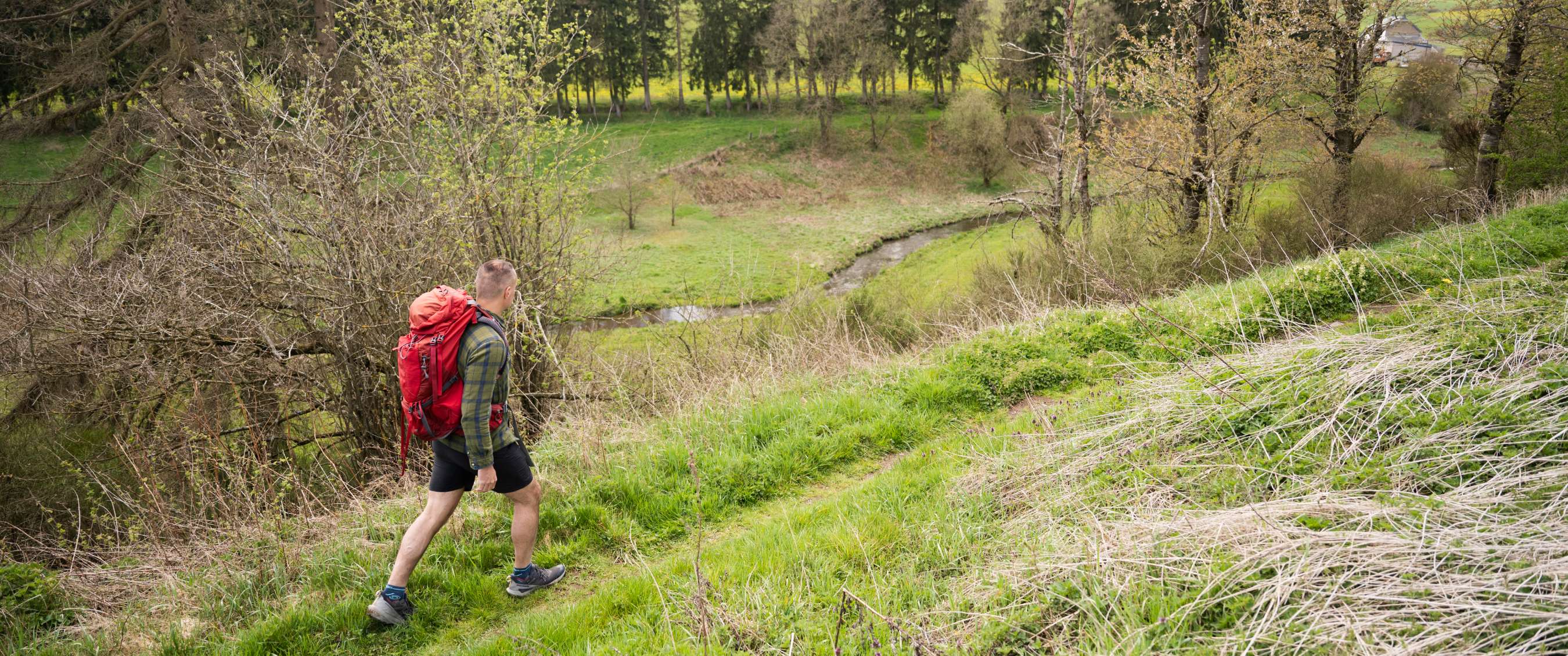 Persoon met rugzak wandelt over gras langs een beek in een heuvelachtig landschap.