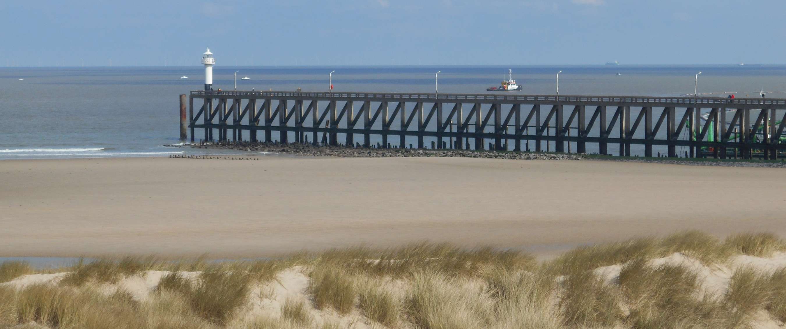 Lange pier met vuurtoren die uitloopt in zee, gezien vanaf een breed zandstrand met duingras.