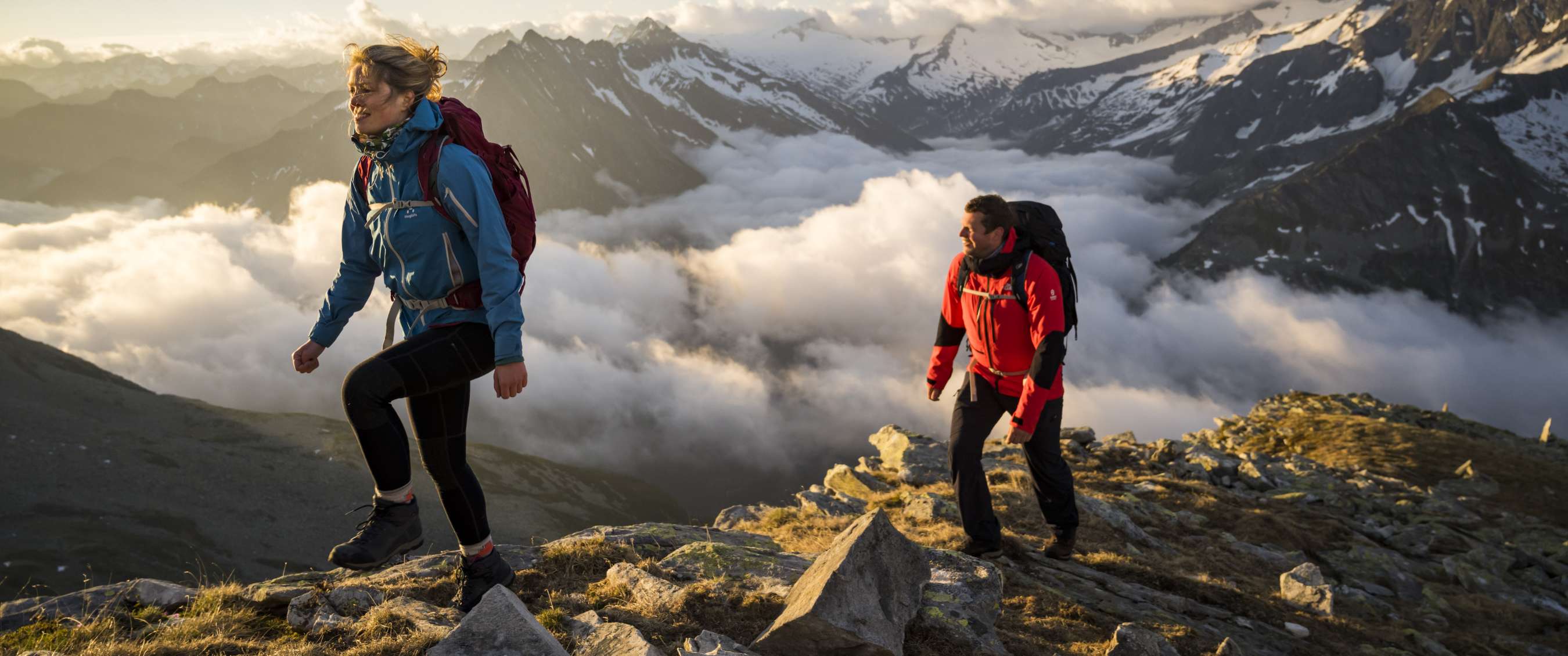 Twee wandelaars stijgen over rotsachtig bergpad boven wolken en sneeuwtoppen.