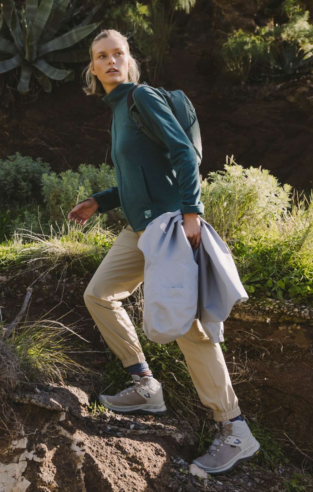 Vrouw in beige wandelbroek en blauw fleecevest stapt over een rotsachtig pad met jas in de hand