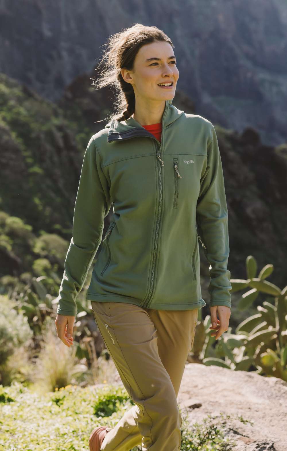 Vrouw in groene jas en beige wandelbroek wandelt door een zonnig berglandschap
