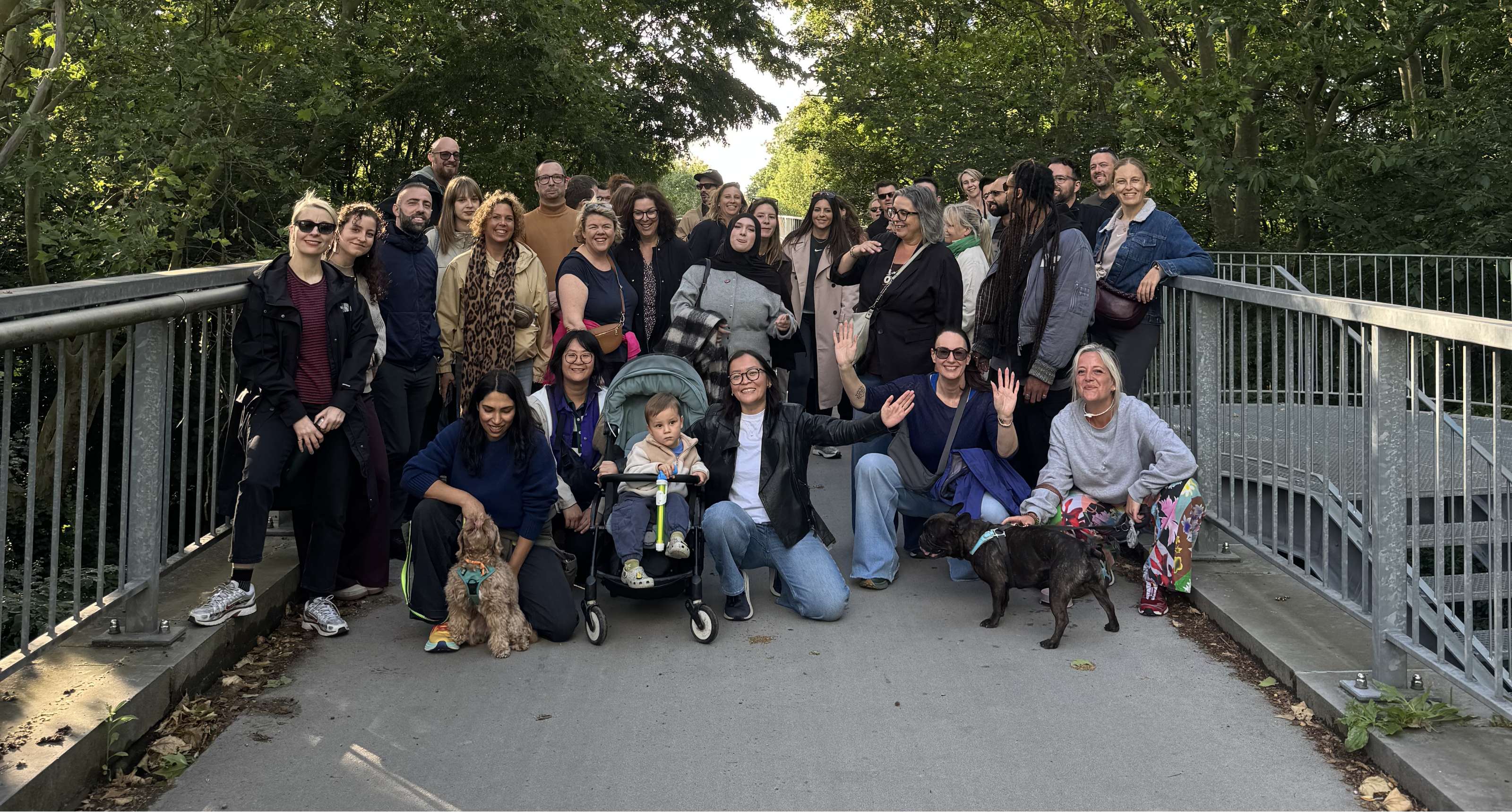 Een groep wandelaars poseert op een brug tussen de bomen, met twee honden en een kind in een buggy.