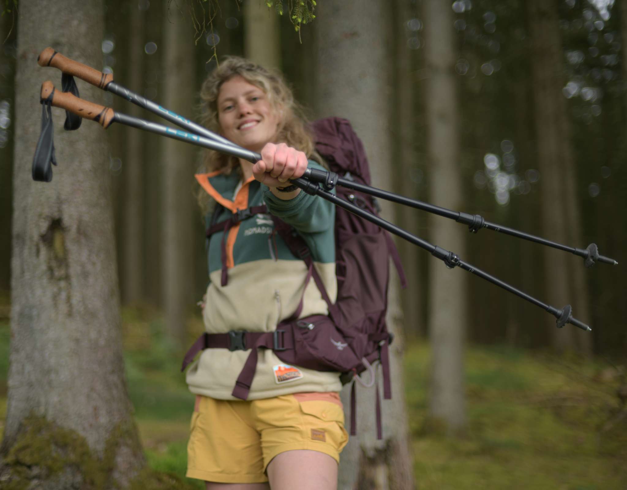 Vrouw met rugzak in het bos houdt glimlachend twee wandelstokken naar voren.