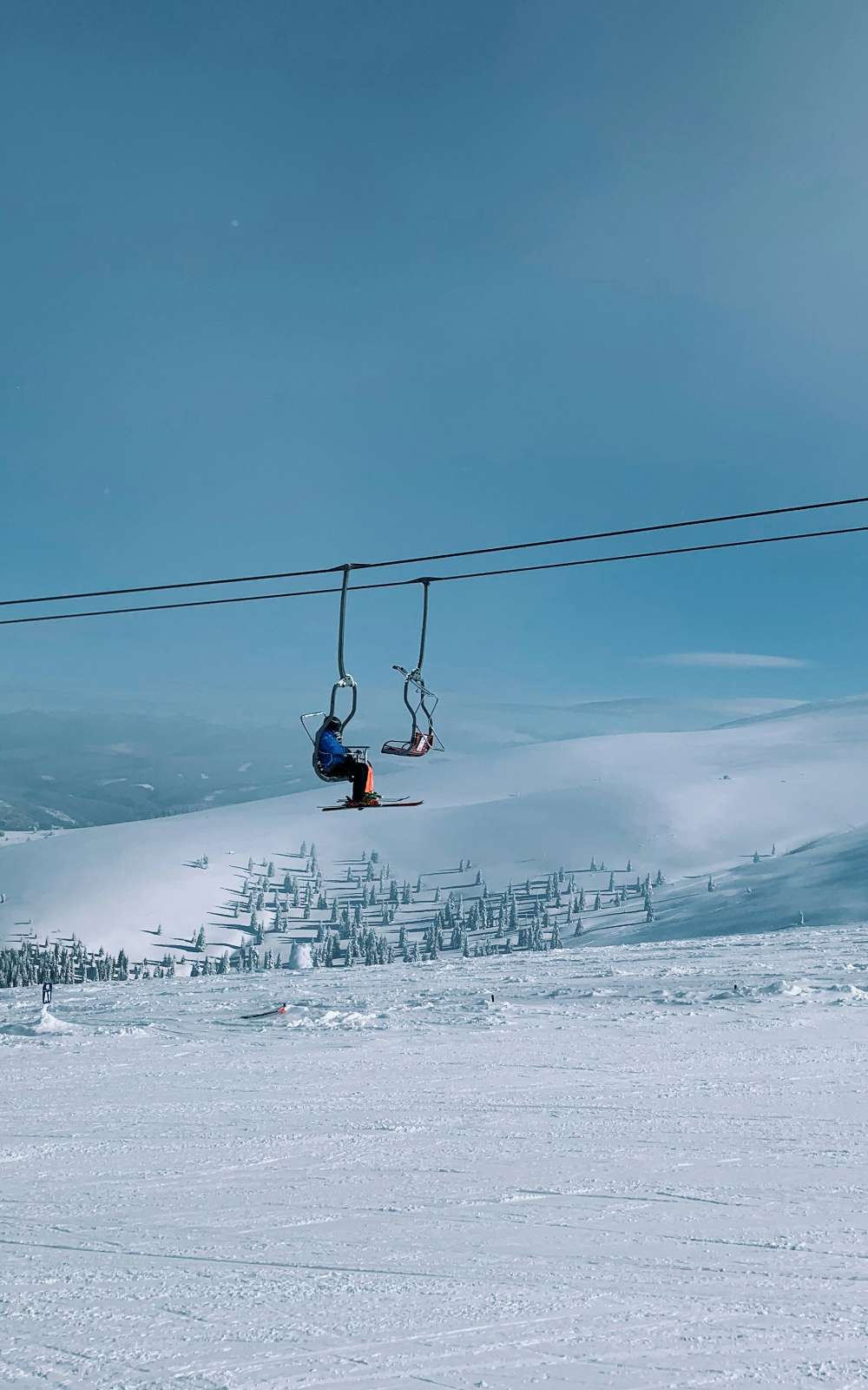 Skiër in een stoeltjeslift boven een besneeuwde helling met naaldbomen.