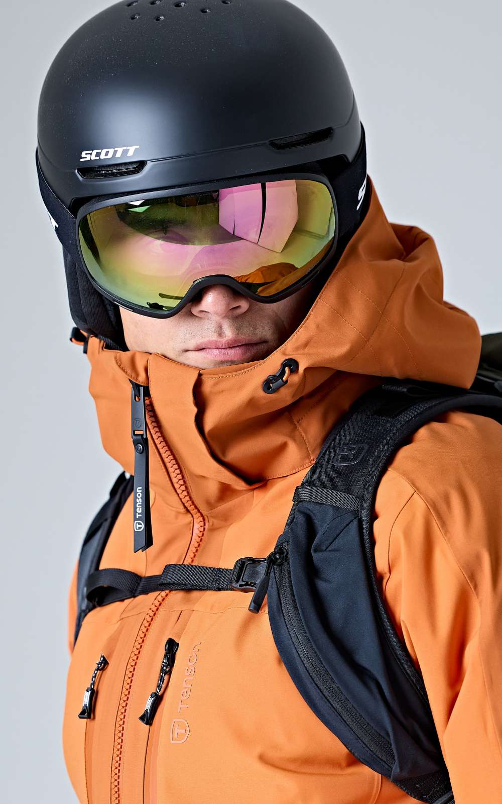 Persoon met oranje ski-jas, zwarte helm, spiegelende skibril en rugzak.