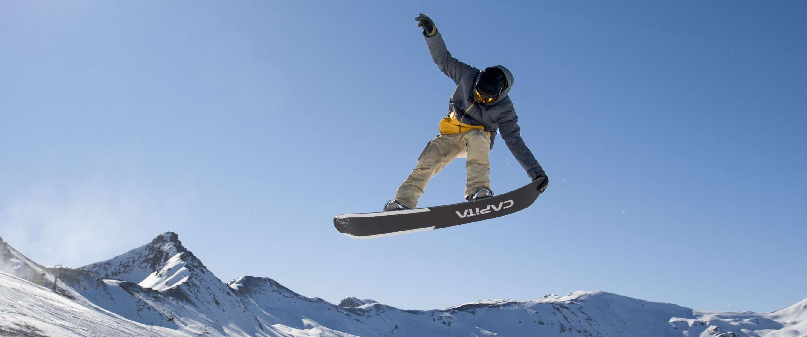 Snowboarder maakt een sprong boven de piste met besneeuwde bergen op de achtergrond.