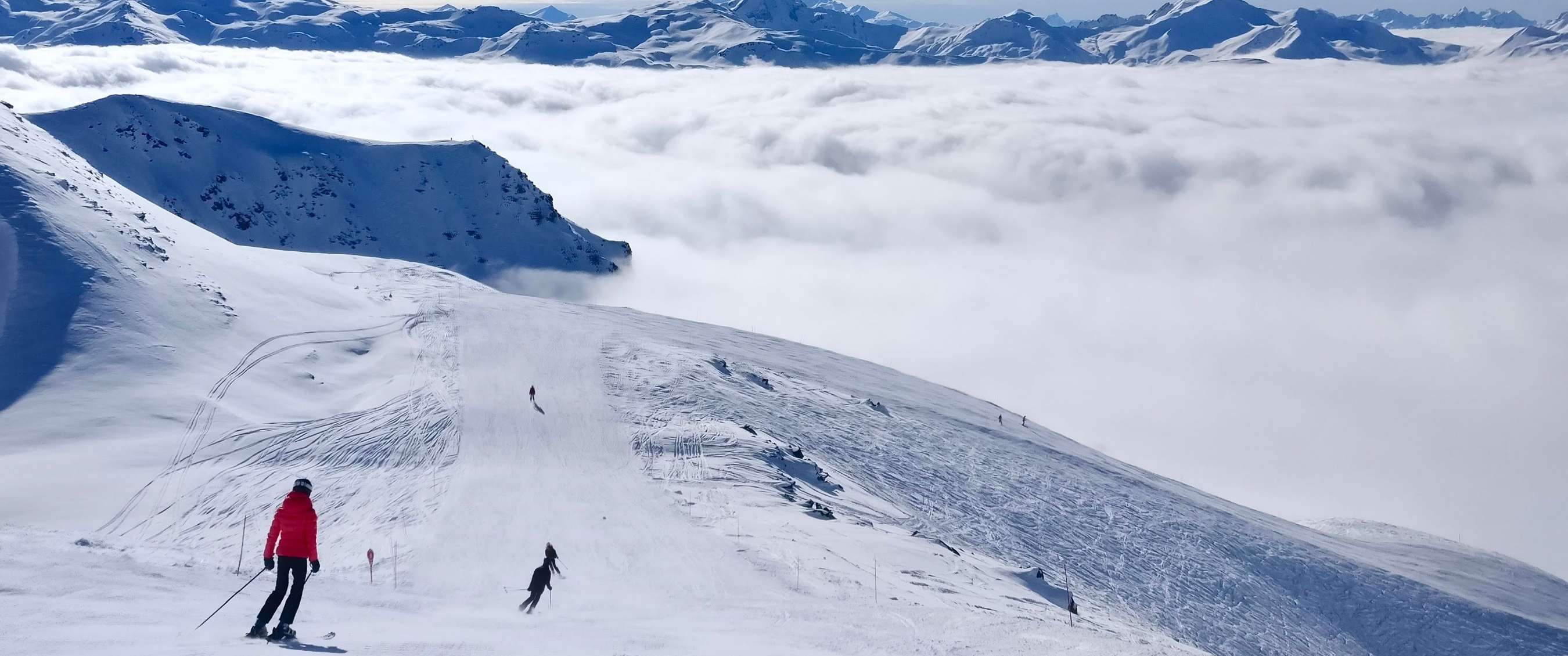 Skiërs dalen een besneeuwde piste af boven een uitgestrekt wolkendek.